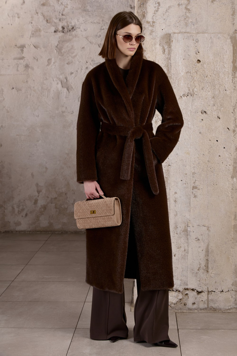 A woman wears this longline coat featuring oversized pockets perfect for sinking hands into, with a generous collar and lapels that create a bold statement. She styles it with the matching thick faux fur belt, cinching the waist for a flattering silhouett