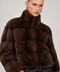 A model wearing a cropped, dark brown fur jacket with a high collar and black leather pants, photographed against a neutral light gray studio background.